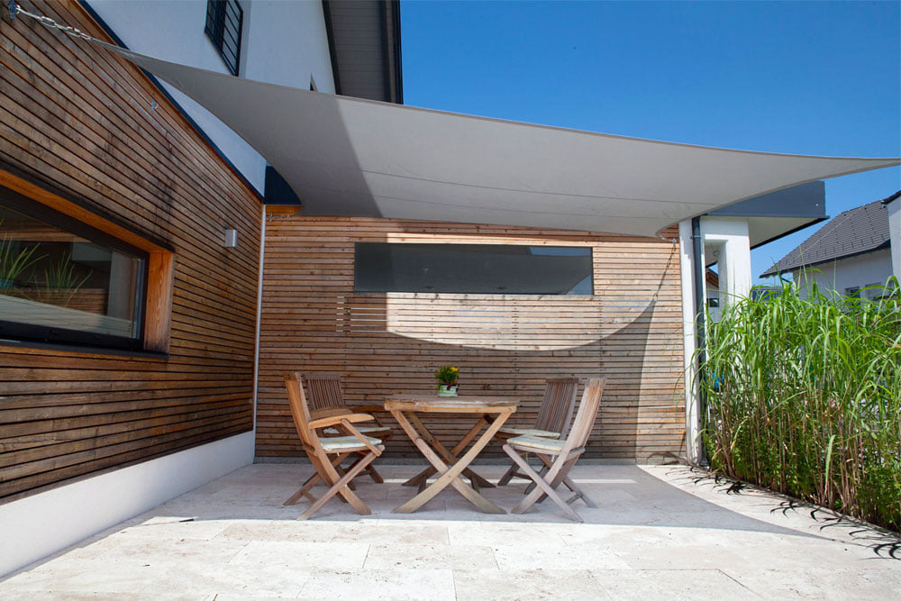 Toldo vela tensado sobre un comedor exterior junto a una fachada de madera